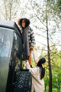 Side view of young african american woman helping friend climbing up camper van parked in green forest during summer adventure in countryside