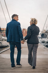 Full length rear view of senior couple holding hands while walking on pier at harbor