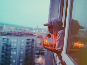 Side view of young man with illuminated cigarette lighter carrying rose in mouth while leaning on window