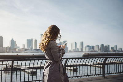 Side view of woman using smart phone on footpath by harbor in city