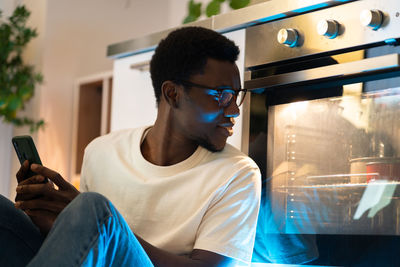 African man sitting near over in kitchen, waiting for cake to bake, cooking sweet surprise for wife