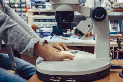 Unrecognizable kid using microscope while examining circuit board in engineering lab.