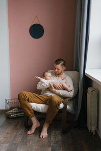 Portrait of a caring caucasian dad holding a cute baby son in his arms, sitting in a comfortable