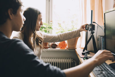 Teenage girl adjusting smart phone on tripod while sitting with friend by computer at home