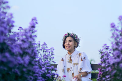 Portrait of smiling young woman standing against purple flowering plants against sky