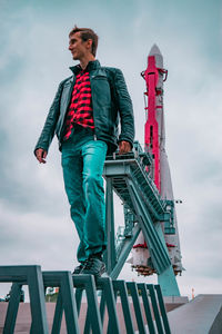 Low angle view of young man looking away while standing on railing against sky