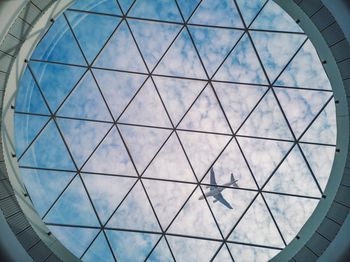 Low angle view of airplane flying against cloudy sky seen through skylight in building