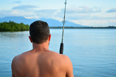 Rear view of shirtless man fishing in lake against sky