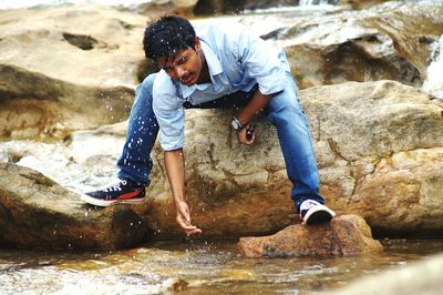 Full length of man washing face white sitting on rock at riverbank