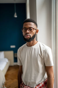 Portrait serious african american bearded man in eyeglasses in white t-shirt near window at home