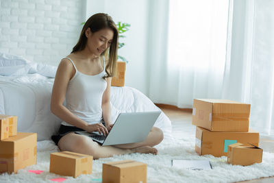Full length of woman using laptop while sitting amidst cardboard boxes at home