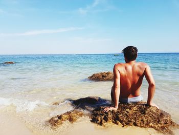 Rear view of shirtless man sitting on rock against sea