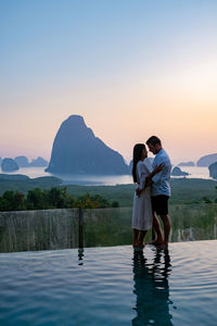 Rear view of couple standing on mountain against sky during sunset