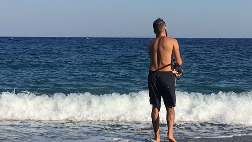 Rear view of shirtless man standing at beach against clear sky