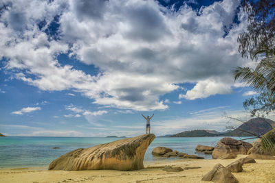 Distant view of man standing on rock formation by sea against cloudy sky