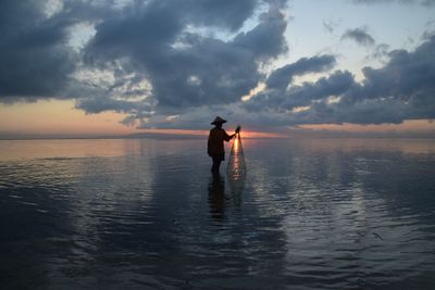 Silhouette fisherman holding fishing net on sea against sky during sunset
