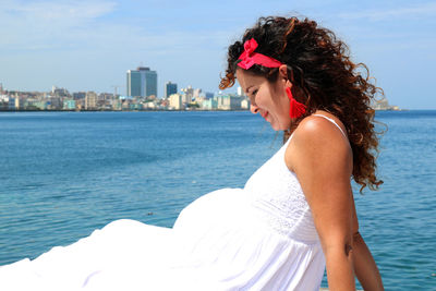 Young pregnant woman dressed in white sitting relaxed by the seaside, looking at her belly 