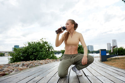 Side view of young man drinking water while sitting on boardwalk against sky