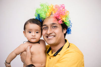 Portrait of smiling father wearing wig carrying baby girl against wall