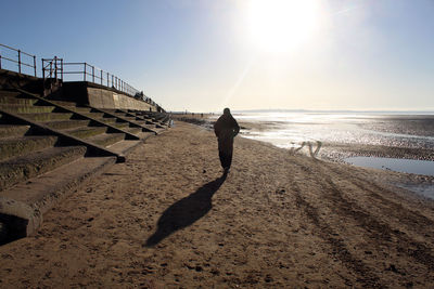 Rear view of man walking at beach against sky during sunset