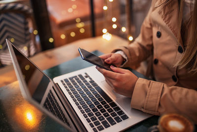 High angle of cropped unrecognizable young female freelancer messaging on mobile phone while sitting at table with laptop and cup of coffee in cozy cafe and working or remote project