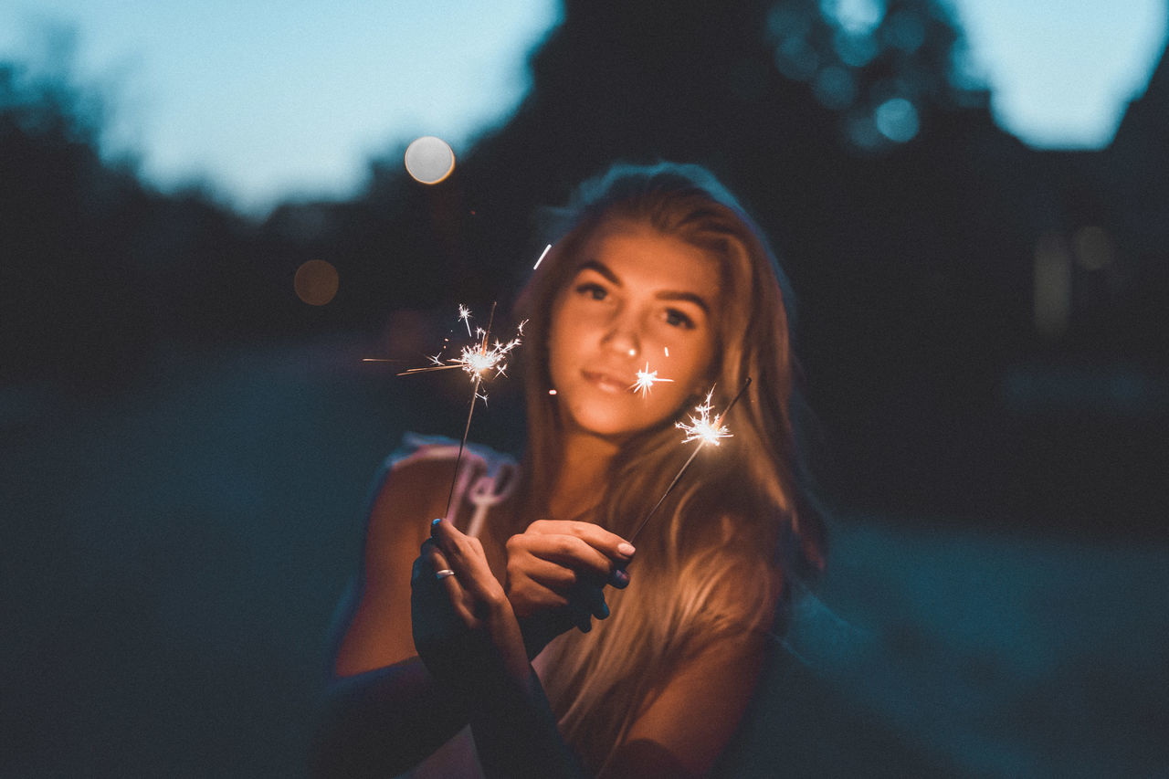 one person, illuminated, real people, night, holding, glowing, leisure activity, front view, portrait, sparkler, lifestyles, focus on foreground, looking at camera, young adult, young women, women, burning, firework, adult, smiling, sparks, firework - man made object, outdoors, hairstyle, beautiful woman