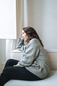 Thinking woman forty year with brunette long hair in cozy knitted sweater near window at home