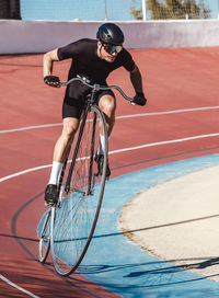 From above adult focused cyclist in black sportswear and helmet riding penny farthing along racetrack at sports stadium while getting ready for competition in sunny day