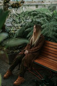 Thoughtful young woman sitting on bench against plants