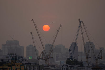 Low angle view of cranes and buildings against sky during sunset