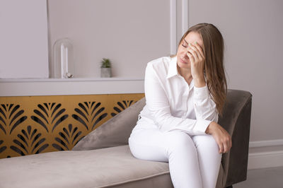 A tired woman after work sits on the couch and closes her eyes with her hands