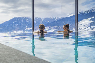 Rear view of female friends swimming in pool against snowcapped mountains