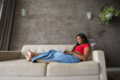 Young black woman working at home  laptop on her lap lying down on  couch. home office gray notebook 