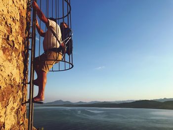 Side view of man with baby boy climbing ladder on wall against sky