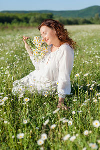 A relaxed woman in a summer loose dress sits among a blooming field of daisies on a clear summer day