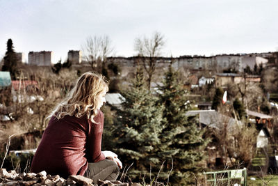 Rear view of young woman looking away while sitting on mountain against sky in town
