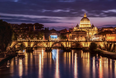 Illuminated bridge over river by buildings against sky at dusk