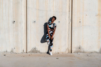 Cheerful confident african american female athlete in flowered sport clothes and white sneakers looking away with interest and laughing while standing alone leaning on concrete wall in sunbeams and resting after training in city