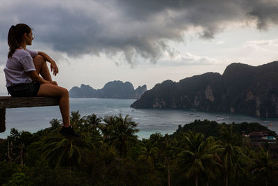 Side view of woman sitting at observation point with sea in background against sky