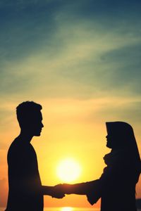 Side view of silhouette couple holding hands while standing against sky during sunset