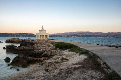 Lighthouse by sea against buildings during sunset