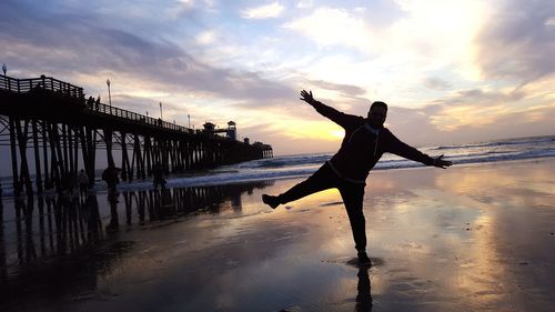 Full length of man with arms outstretched standing at beach against sky during sunset