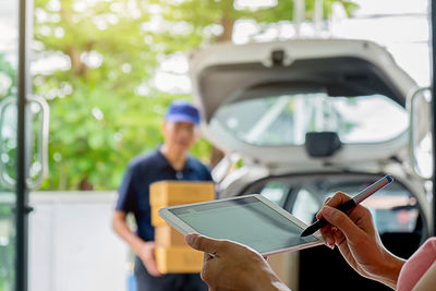 Cropped hands of woman signing on digital tablet with man holding boxes in car