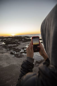 Close-up of man photographing with mobile phone at beach during sunset