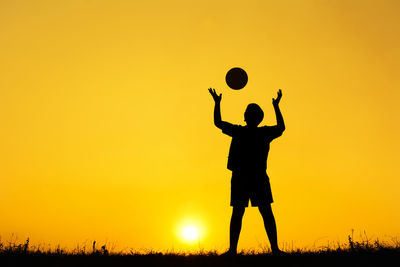 Silhouette boy playing with ball while standing on field against orange sky