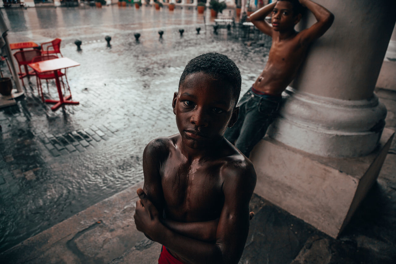 Havana, cuba - december 14, 2019: from above of wet shirtless black fit teen boy feeling cold and looking at camera while standing with friend on city street on rainy day