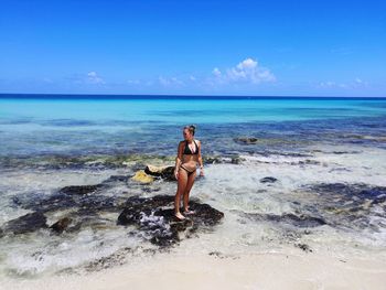 Woman in bikini standing by beach against blue sky