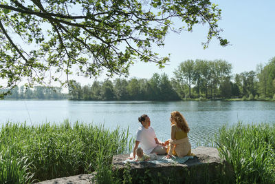 Rear view of friends sitting by lake against trees