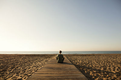Rear view of woman meditating on footpath at beach against clear sky