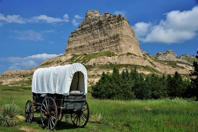 Covered wagon on field against rock formation and blue sky with clouds.